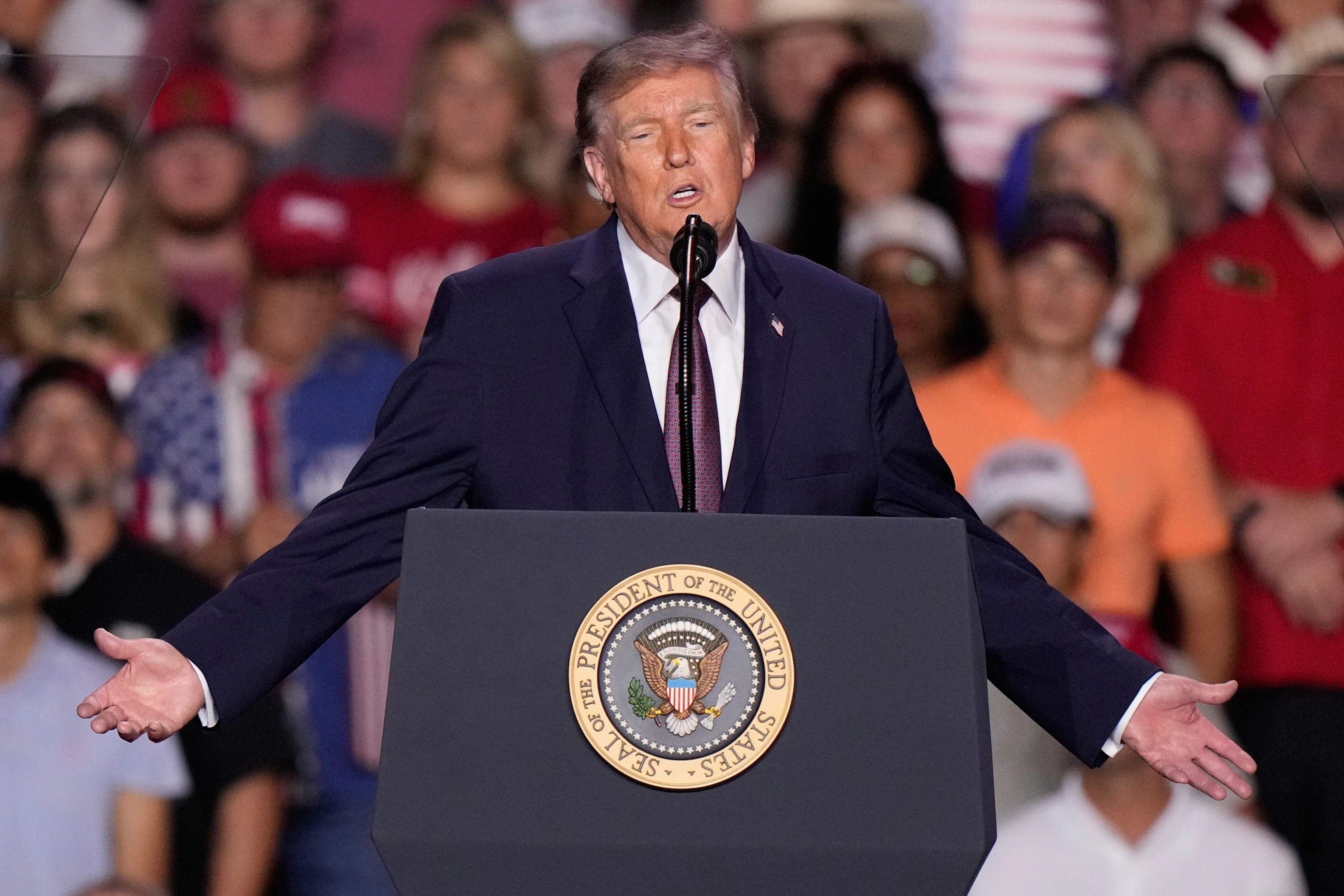Donald Trump speaking from a rally podium, with supporters visible in the background