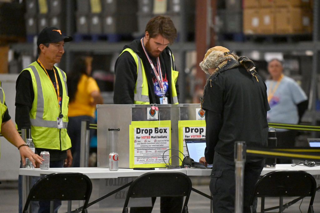 Election workers seated at long tables in a county election office counting ballots under bright overhead lights, documentary news photograph style