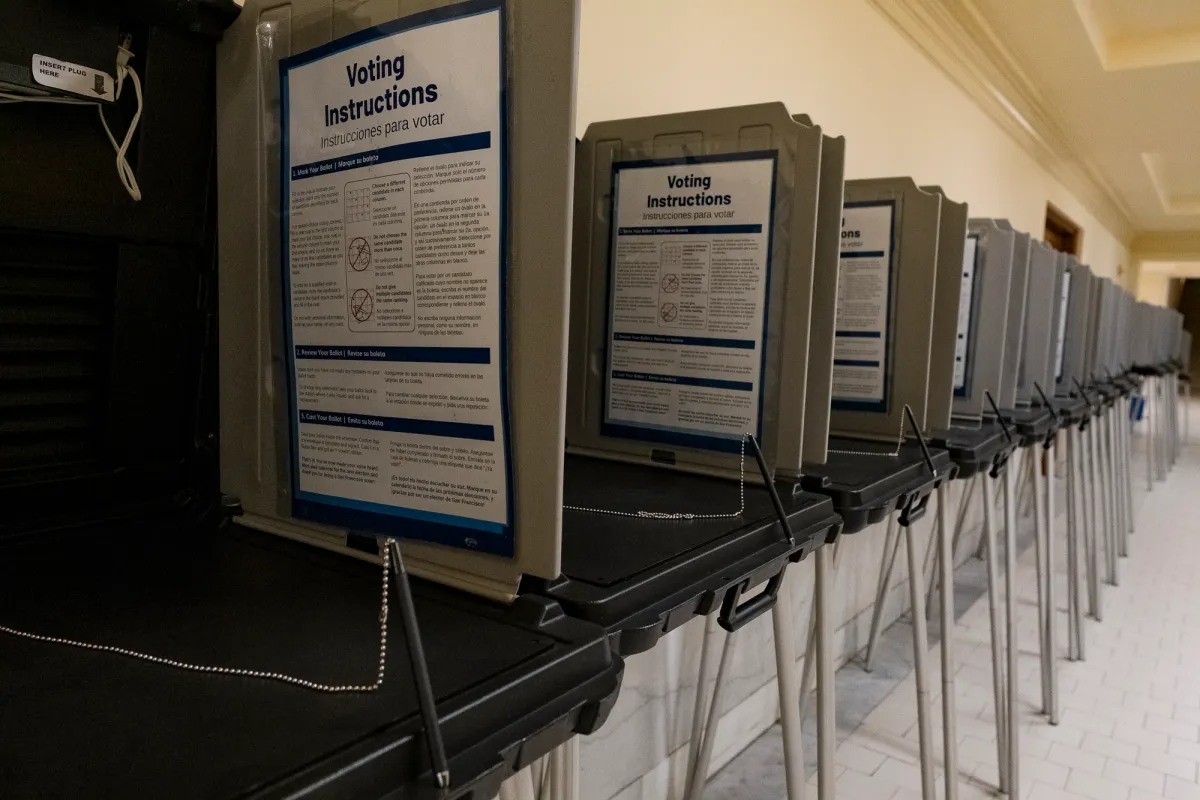Election workers seated at tables in a municipal counting room with stacks of paper ballots and secure ballot containers during a ranked-choice vote tally, realistic news photography style