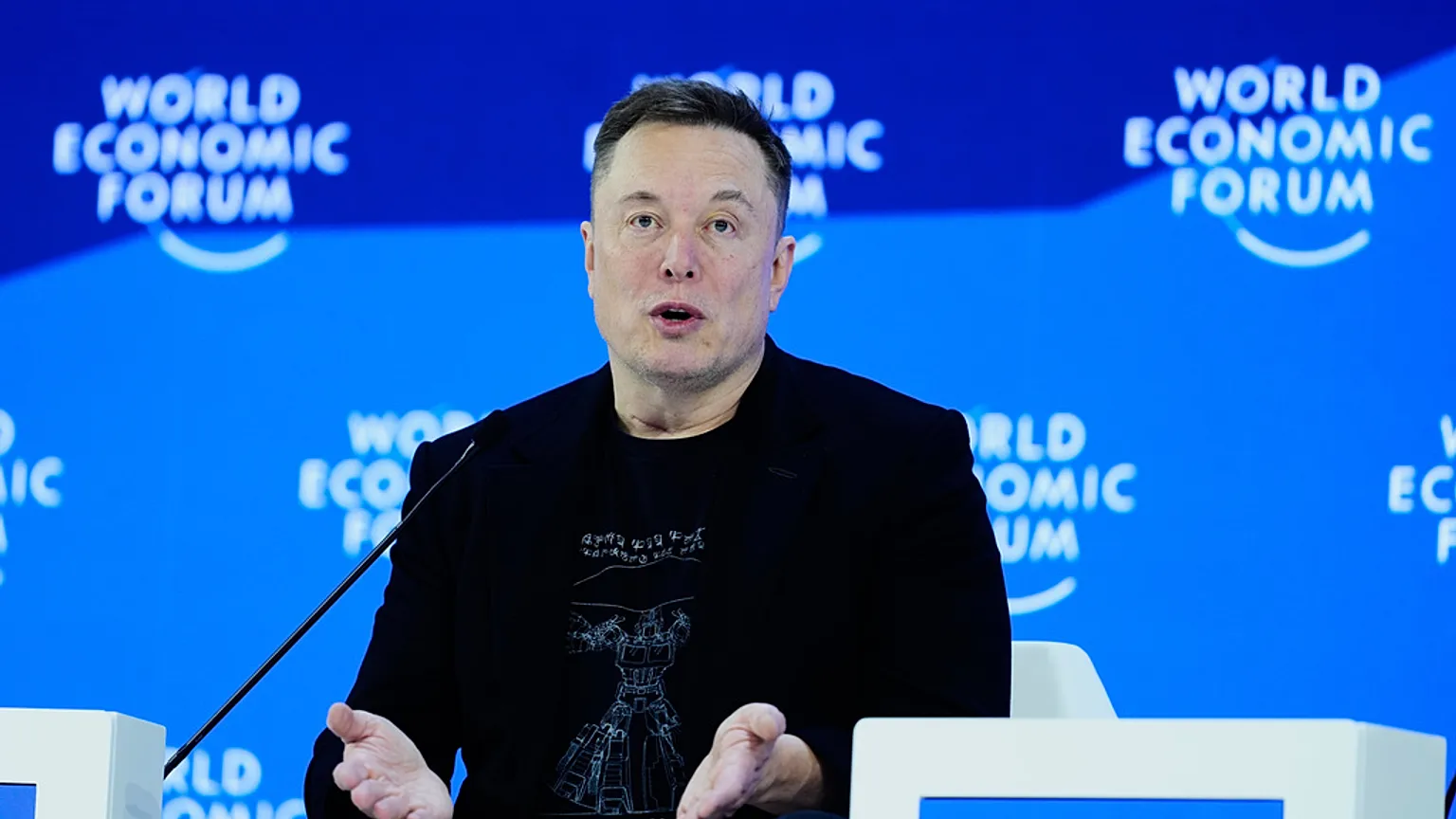 Elon Musk speaking at a public event in 2026, photographed in a candid news style with microphones and a blurred crowd in the background