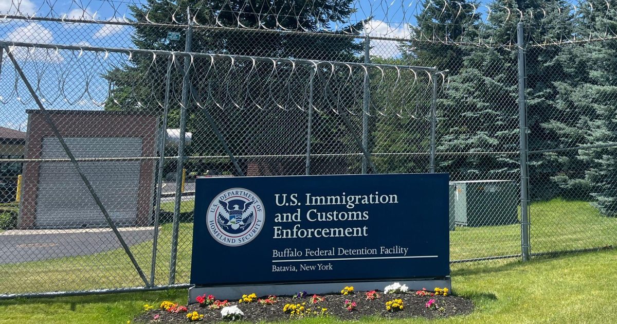 Exterior entrance of a U.S. Immigration and Customs Enforcement detention facility with signage and a secure gate, photographed from a public roadway