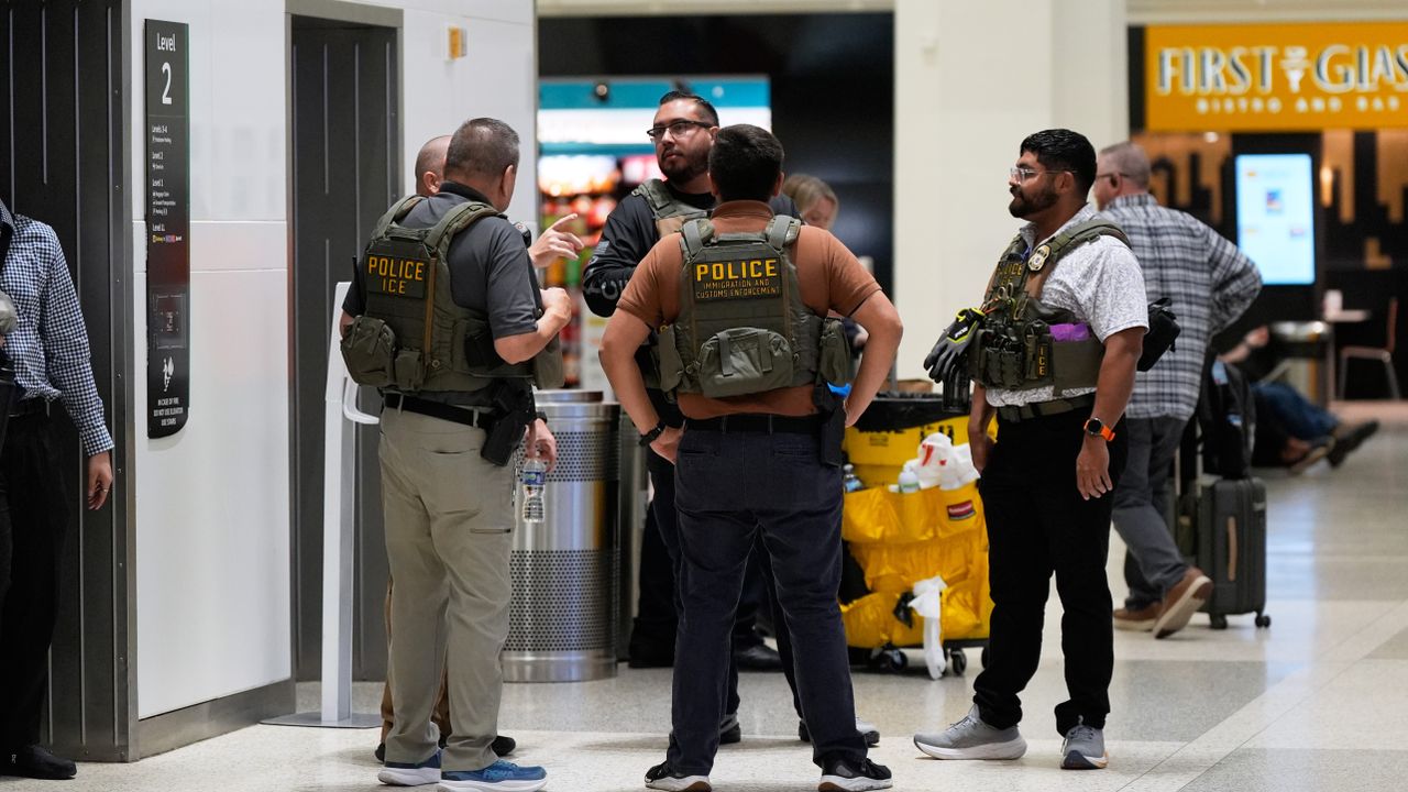 Families and travelers standing in an orderly line inside a United States port of entry inspection area, with officers working at inspection booths in the background, news photography style