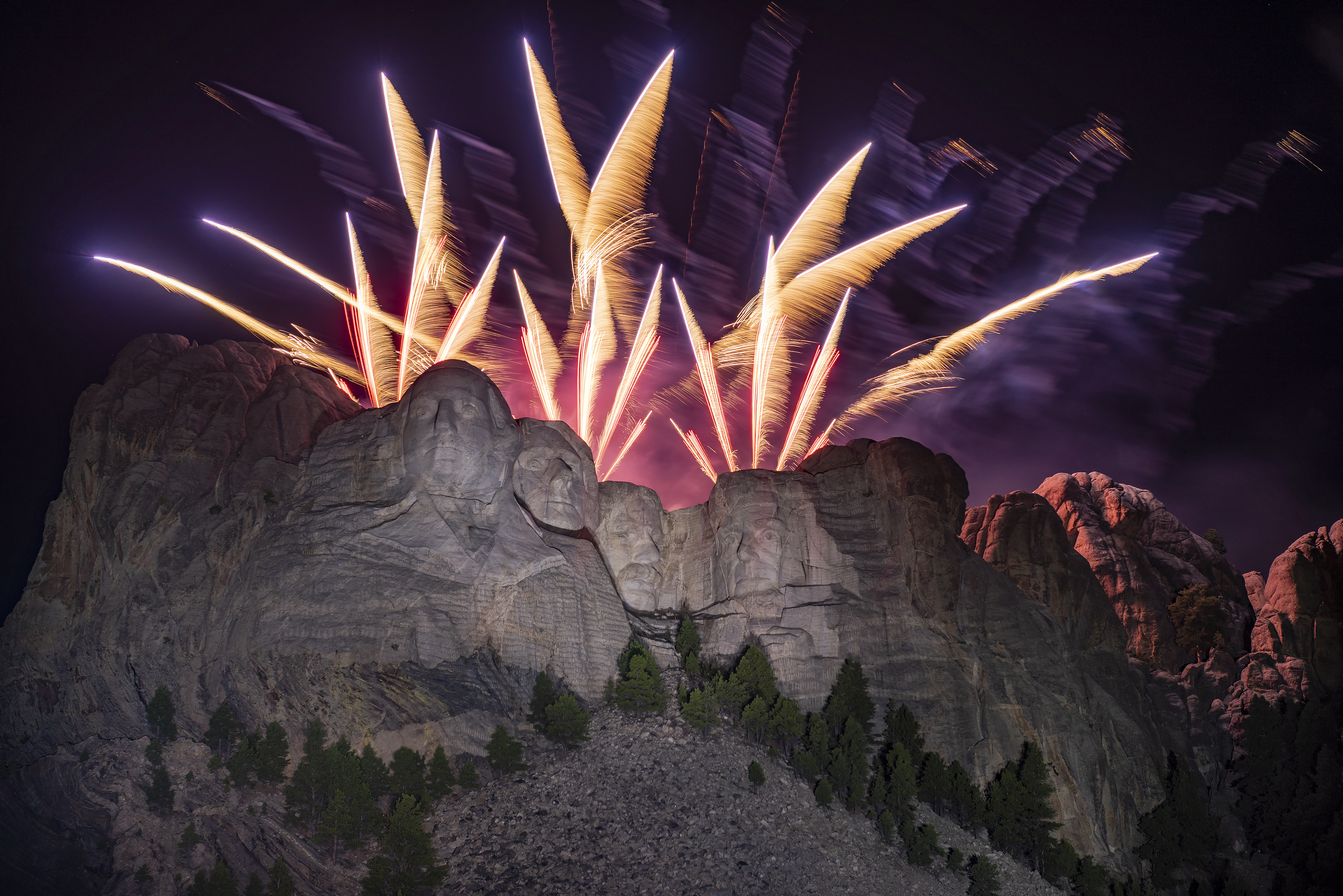 Fireworks bursting above Mount Rushmore at night, with the monument illuminated and spectators watching from the viewing area