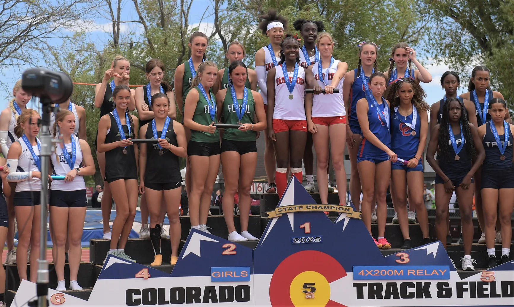 High school girls preparing at the starting line of an outdoor track meet, with officials and spectators in the background, documentary sports photography style
