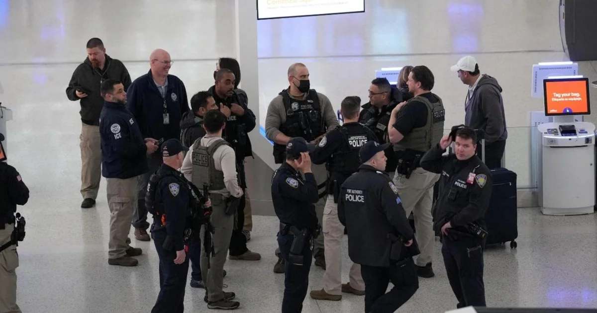 ICE agents walking through a terminal area at John F. Kennedy International Airport in New York City, with travelers and luggage in the background, news photography style