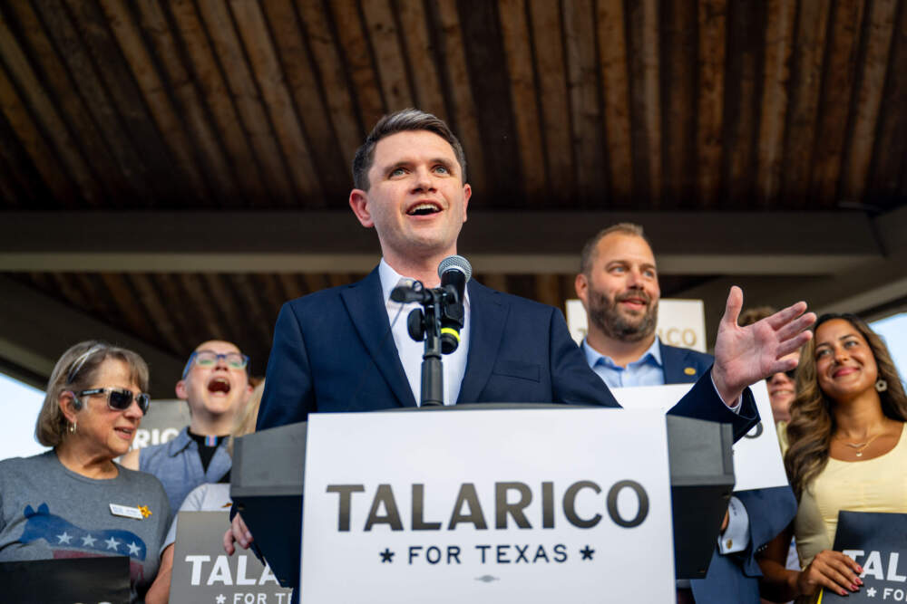 James Talarico speaking at a public campaign event in Texas, standing at a microphone with supporters blurred in the background, photorealistic news photography