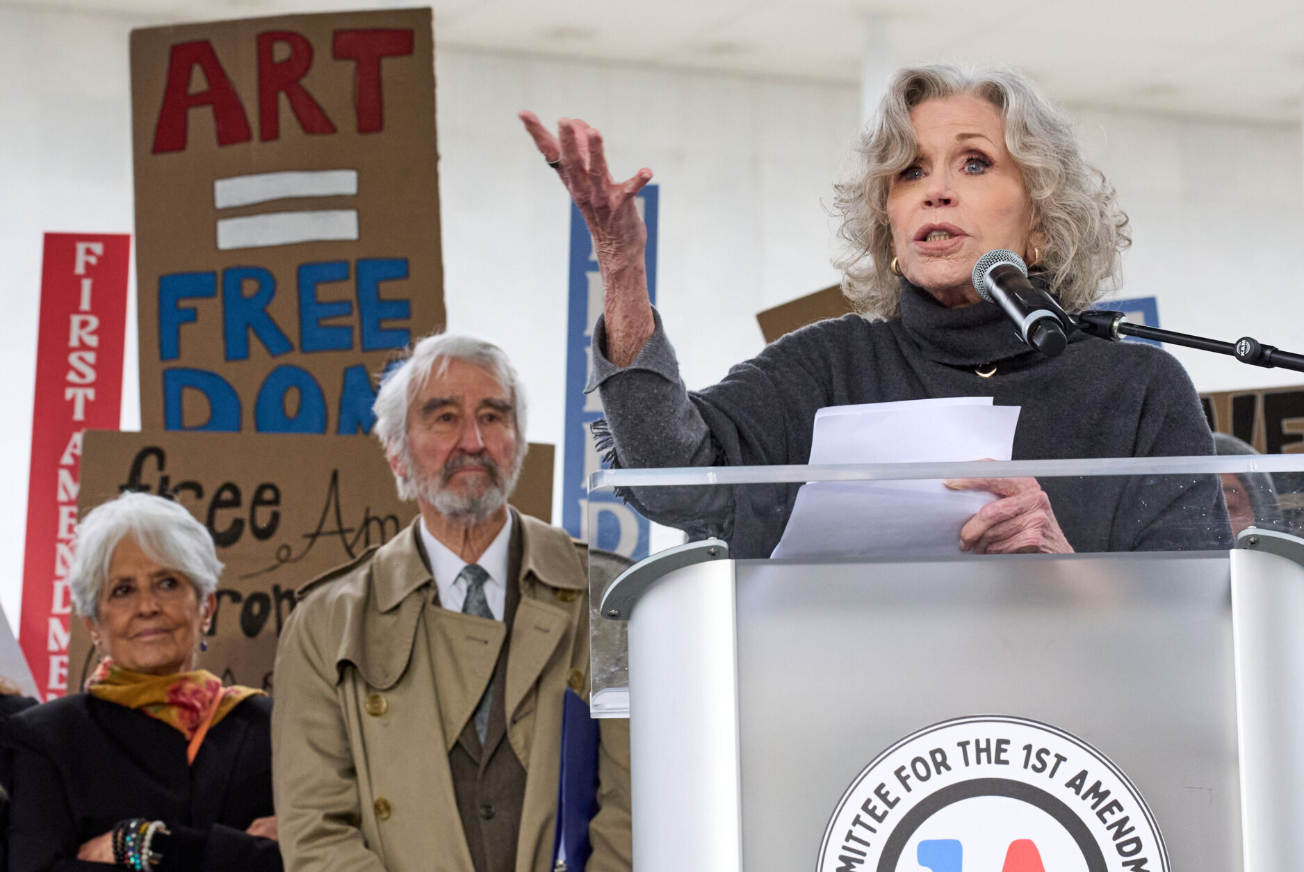 Jane Fonda speaking into a handheld microphone at an outdoor rally near the Kennedy Center, supporters gathered behind her, news photography style