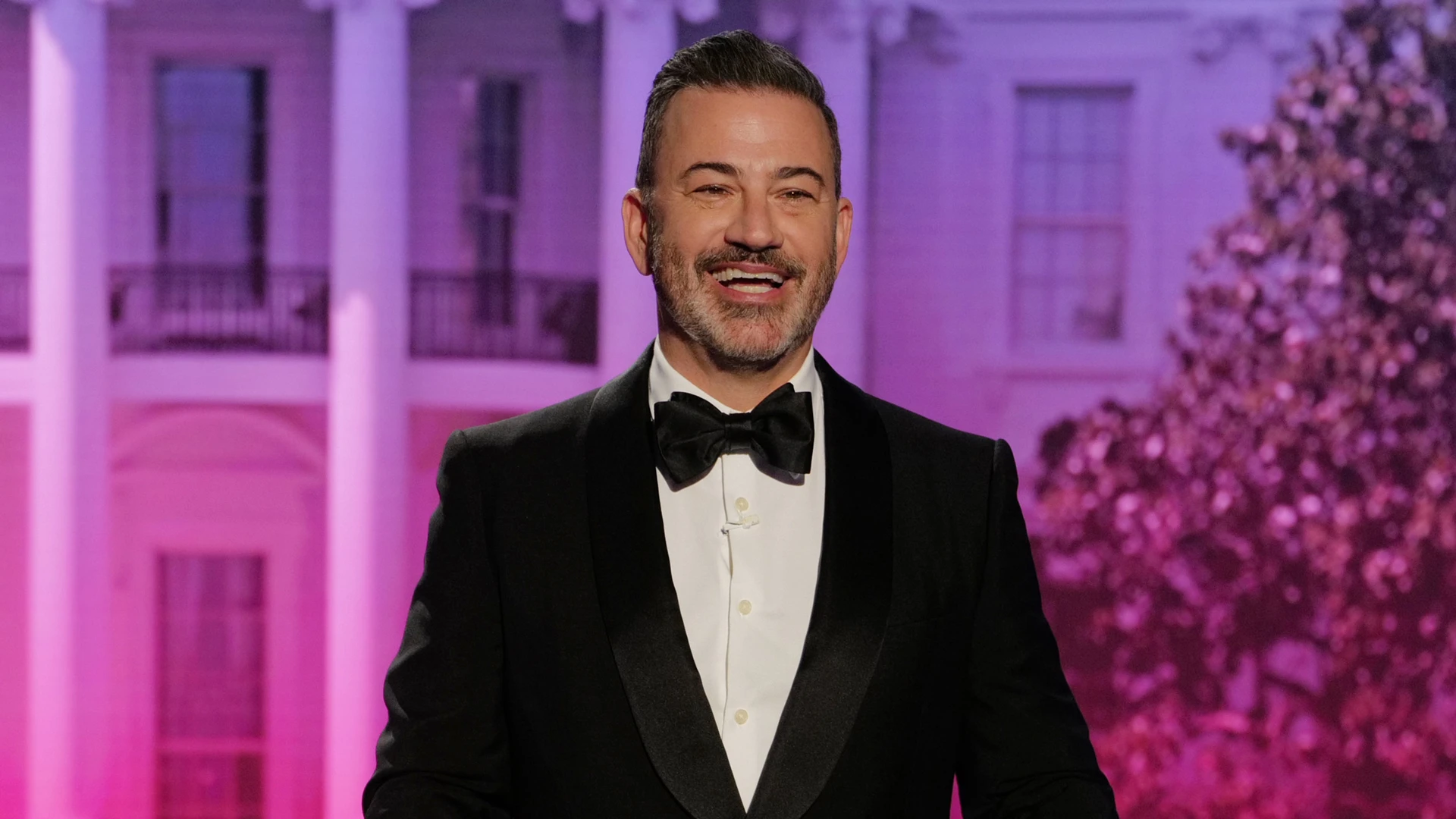 Jimmy Kimmel seated at a desk on a late-night television set with studio lights and audience seating in the background, news photography style