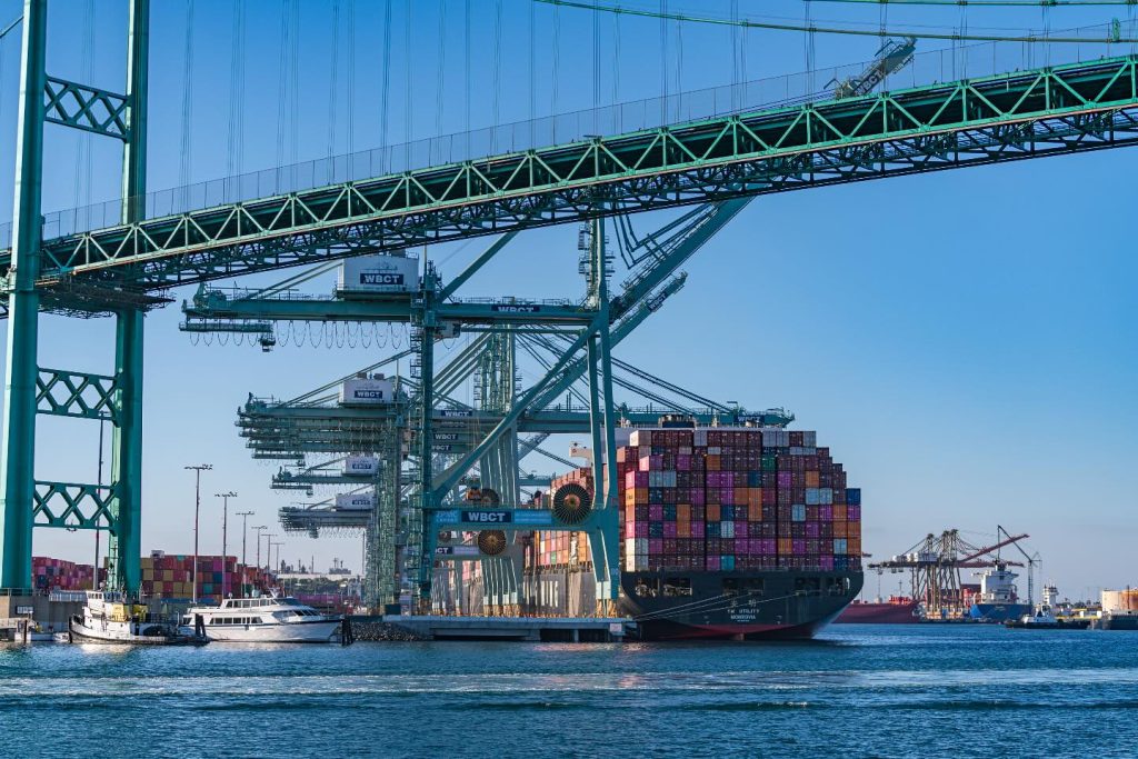 Long rows of cargo containers stacked at the Port of Los Angeles while customs officers inspect shipments near a docked container ship, nighttime news photography style