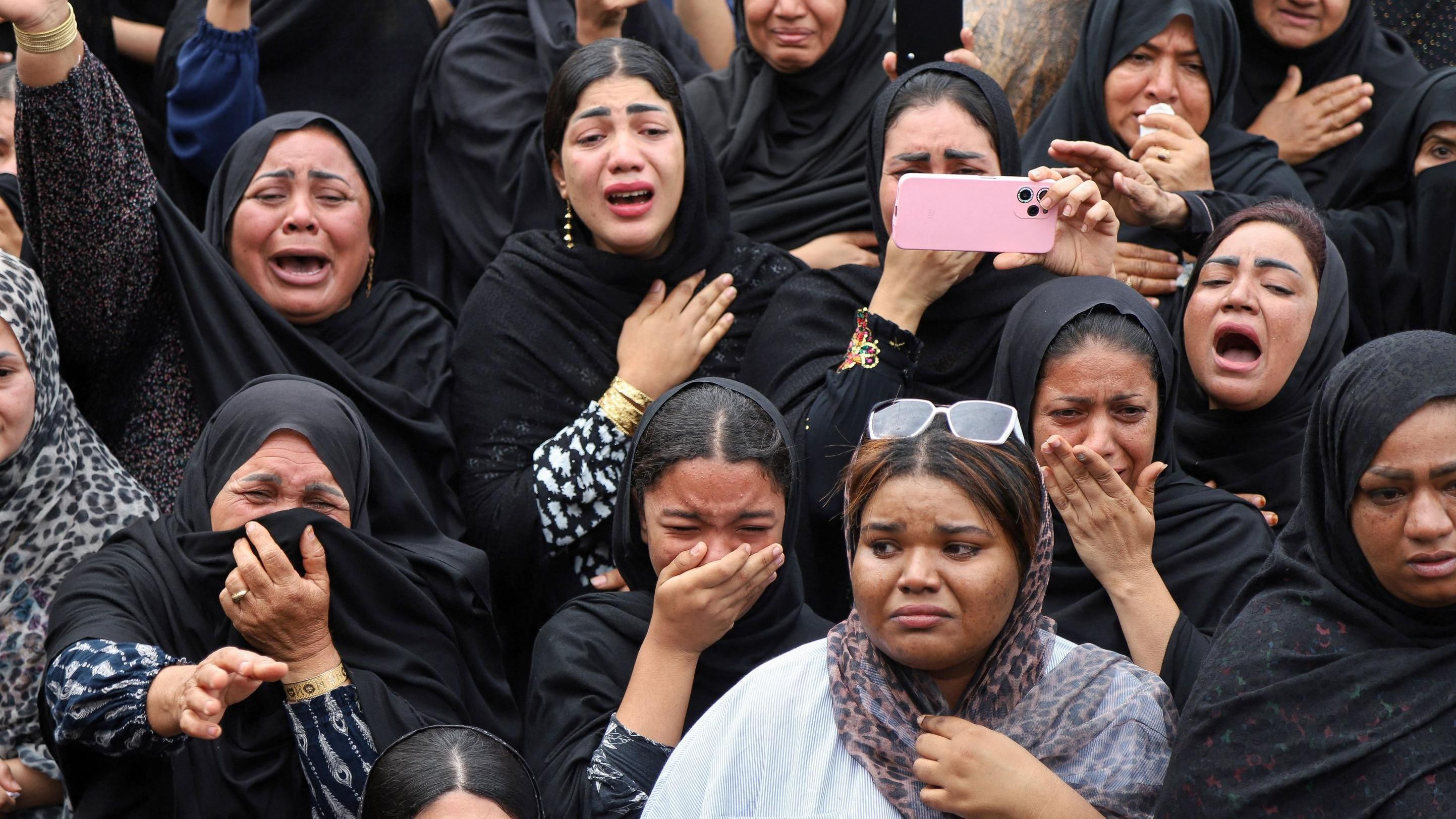 Mourners gathered at a cemetery during a daytime funeral procession, with people standing close together in a somber scene, photographed in a realistic news style