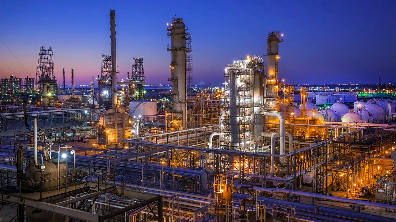 Oil refinery workers in hard hats and high-visibility clothing walking near industrial piping during a shift change, realistic news photography style
