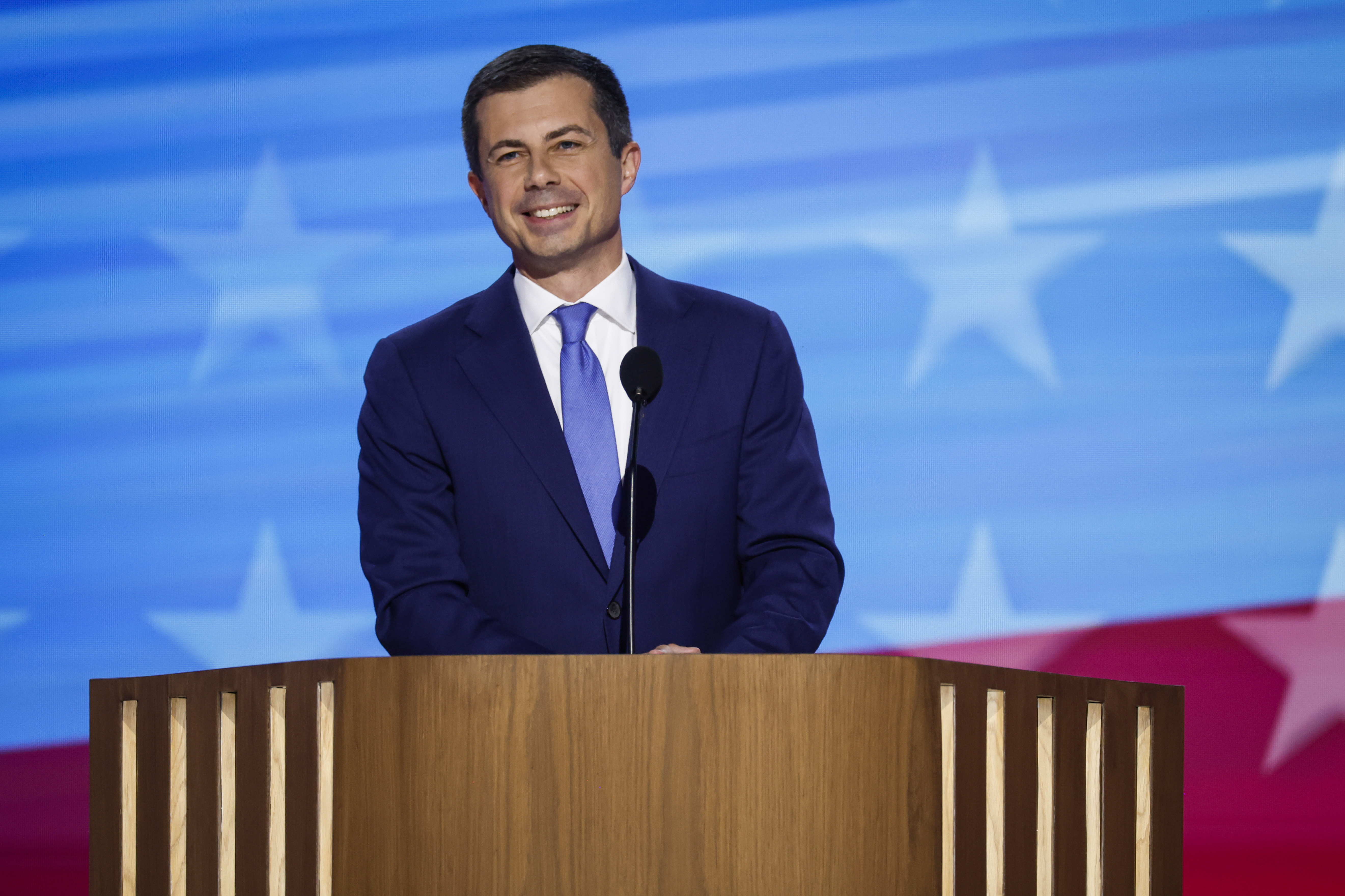 Pete Buttigieg speaking at a public event, standing at a podium with microphones in a candid photograph