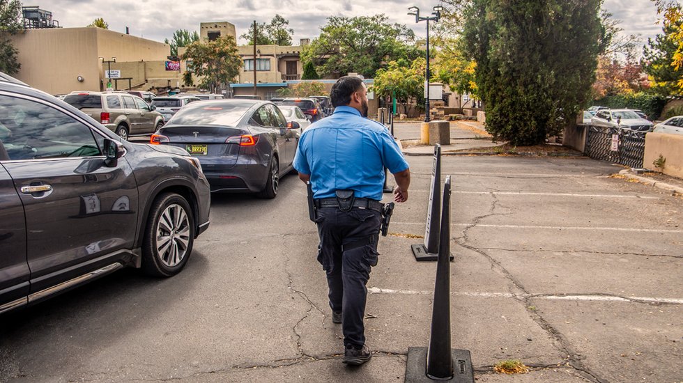 Photograph of a uniformed police officer performing an outer-clothing pat-down on an adult standing with hands visible on a city sidewalk at dusk, with storefront lights in the background