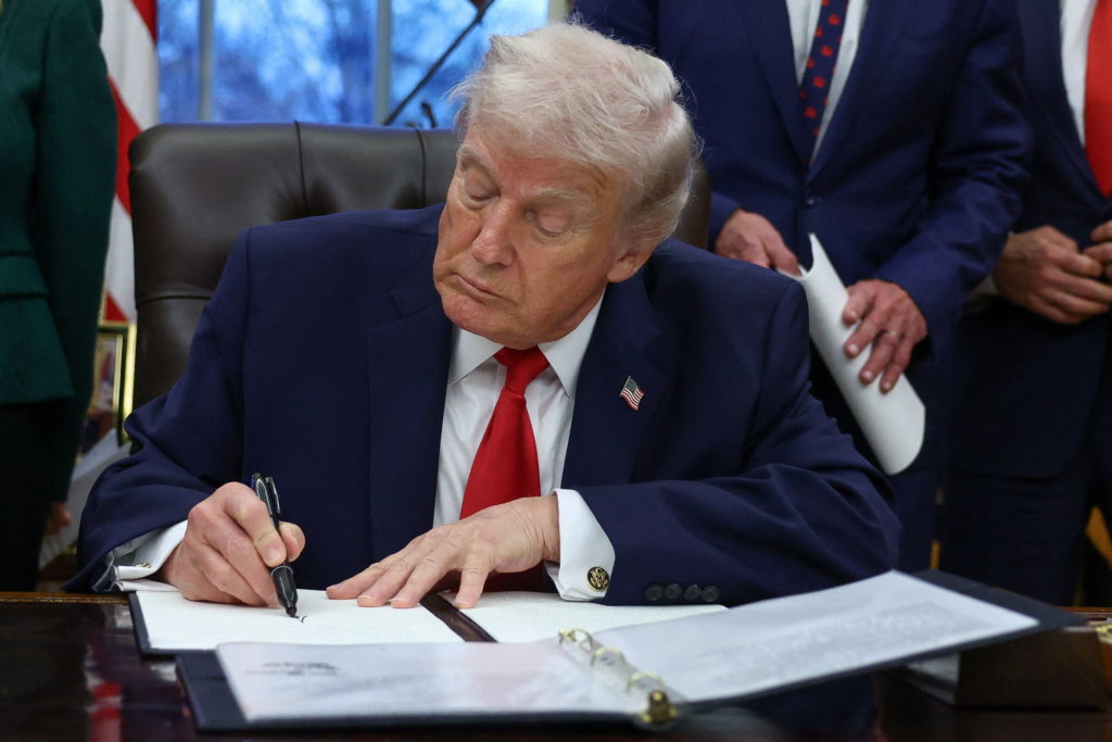President Donald Trump seated at a desk signing an executive order, with staff standing nearby in a formal White House setting, news photography style