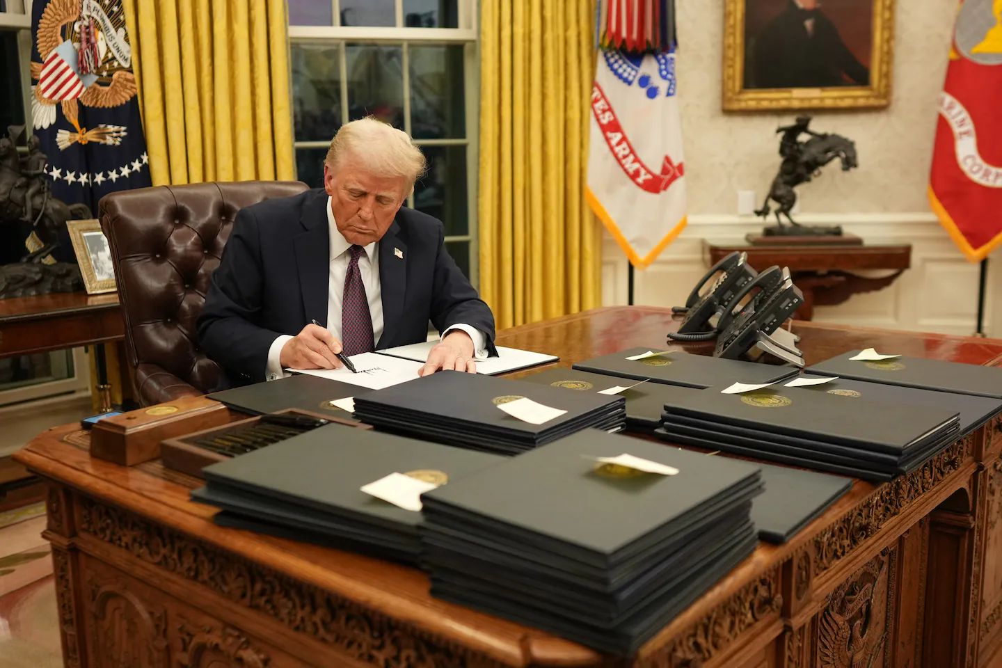 President Donald Trump seated at a desk signing an executive order during a White House event, with staff and photographers nearby
