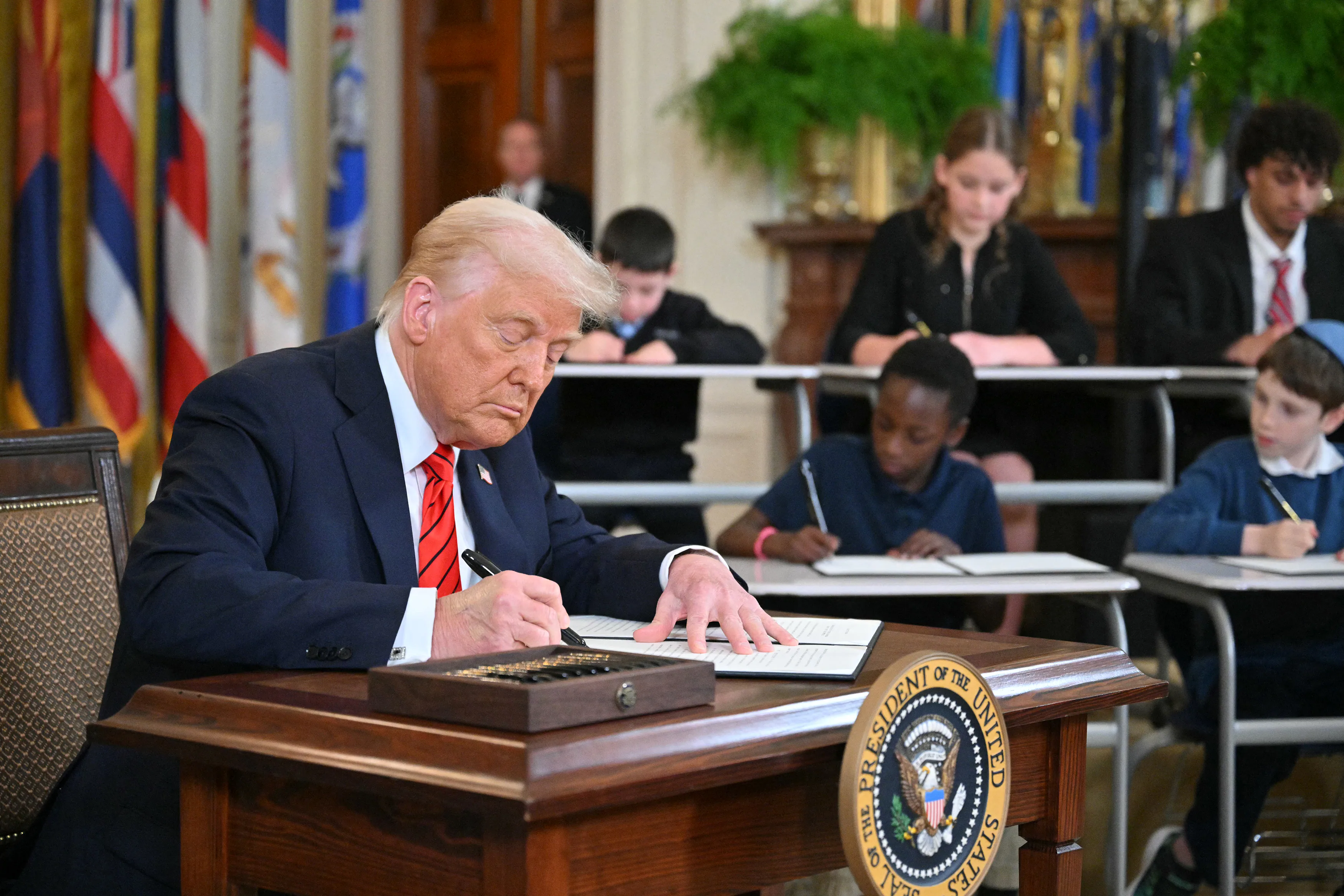 President Donald Trump seated at the Resolute Desk in the Oval Office, holding up a signed executive order with aides and press photographers nearby
