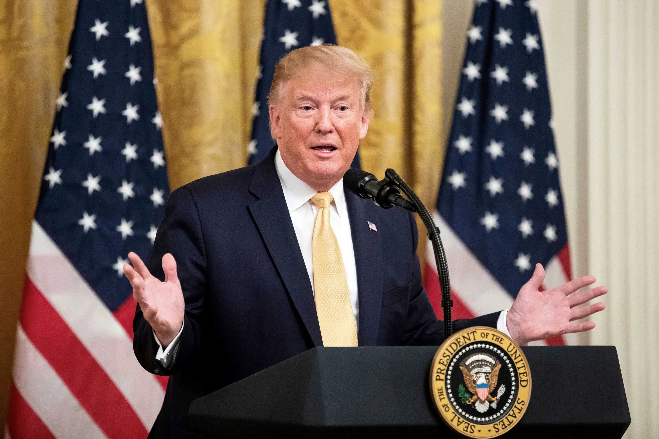 President Donald Trump speaking at a White House event, standing at a lectern with U.S. flags behind him