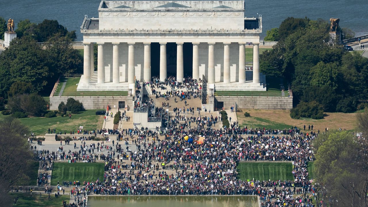 Protesters standing and marching on the National Mall near the Lincoln Memorial in Washington, D.C. on a bright day, with American flags visible, news photography style