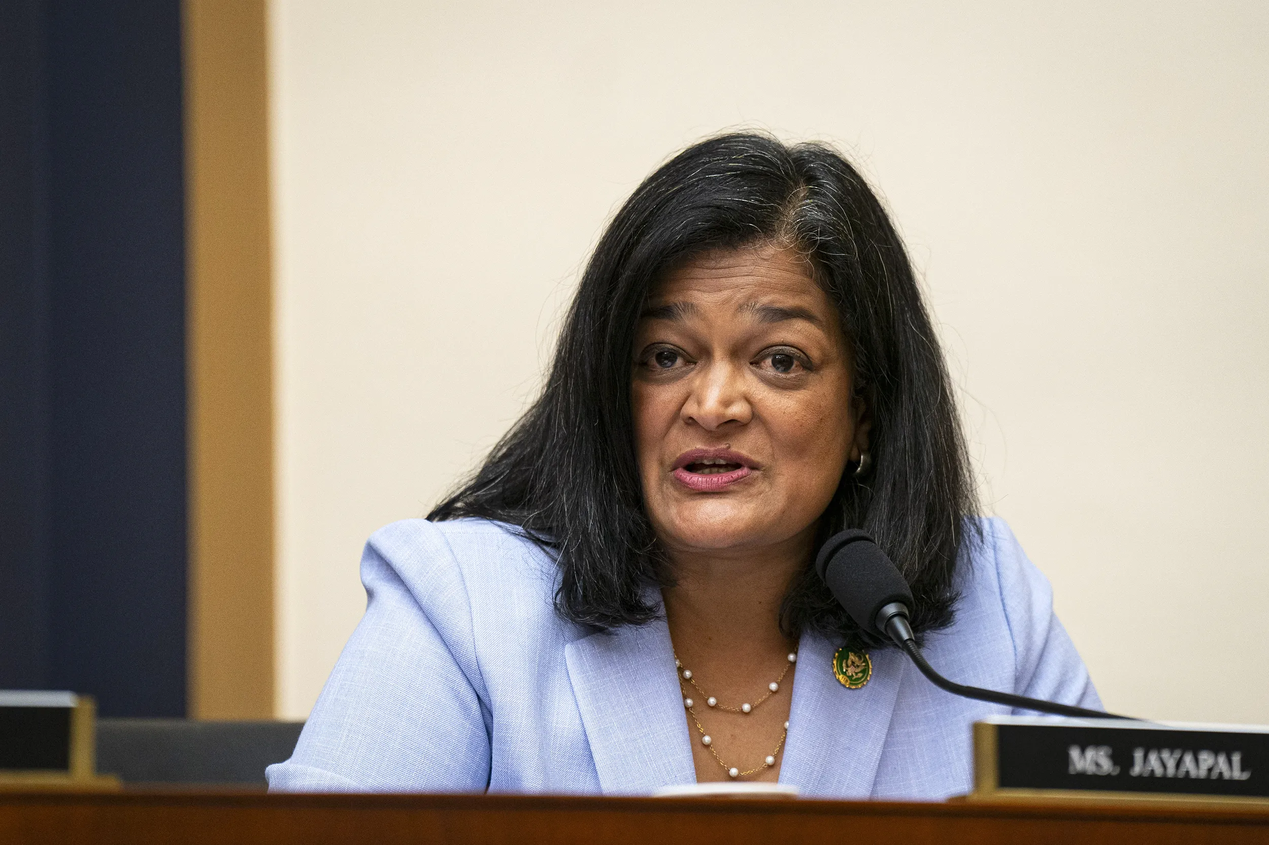 Representative Pramila Jayapal speaking at a congressional-style hearing table with microphones, serious expression, indoor news photo style