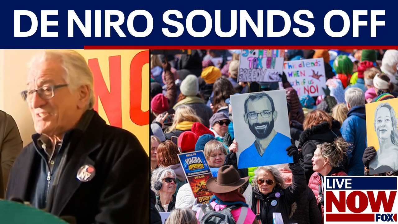 Robert De Niro speaking into a handheld microphone at an outdoor rally with protesters gathered behind him, daytime news photography style