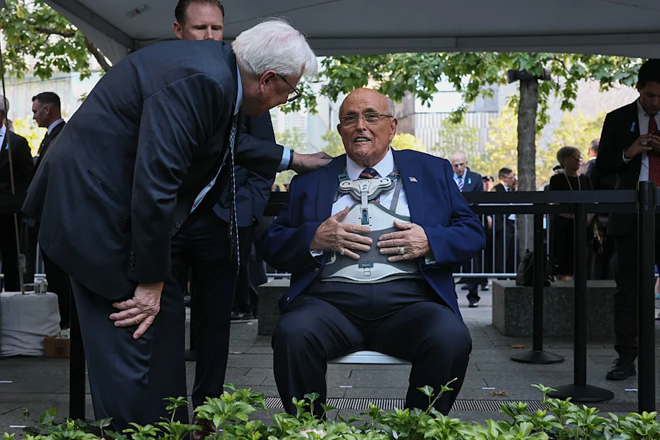 Rudy Giuliani standing in a dark suit at a September 11 anniversary ceremony in New York City, solemn crowd and flags in the background, news photography style