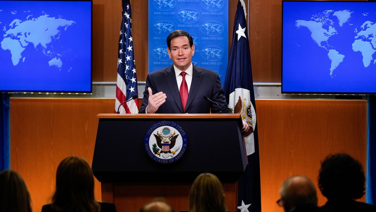 Secretary of State Marco Rubio speaking at a lectern in a U.S. State Department briefing room, with flags behind him, in a real news photograph
