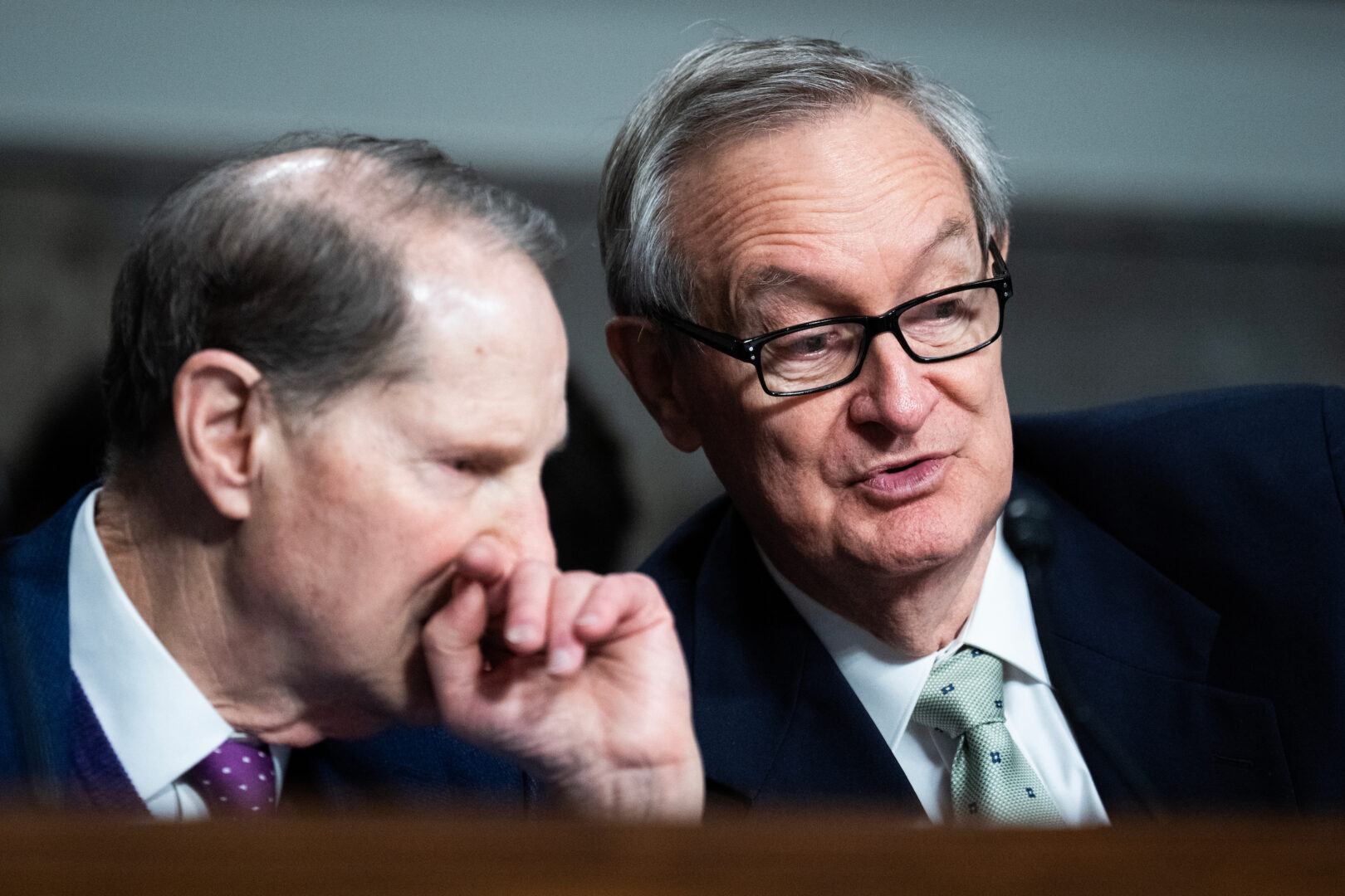 Senator Ron Wyden speaking into a microphone during a Senate committee hearing, papers and nameplate visible, news photography style