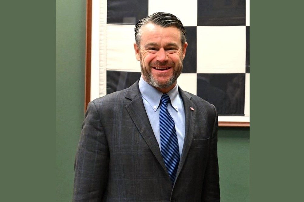 Senator Todd Young speaking at a committee hearing, seated behind a microphone with other senators blurred in the background, candid news photography