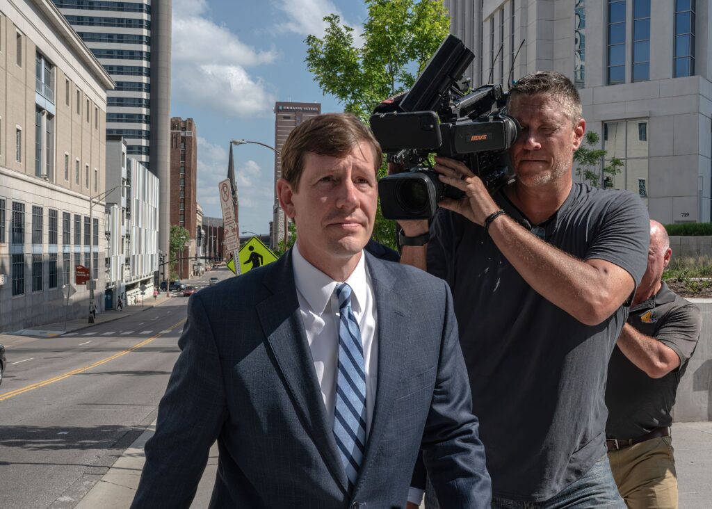 Television news cameras on tripods aimed at the entrance of a federal courthouse as people walk up the steps, real news photography style