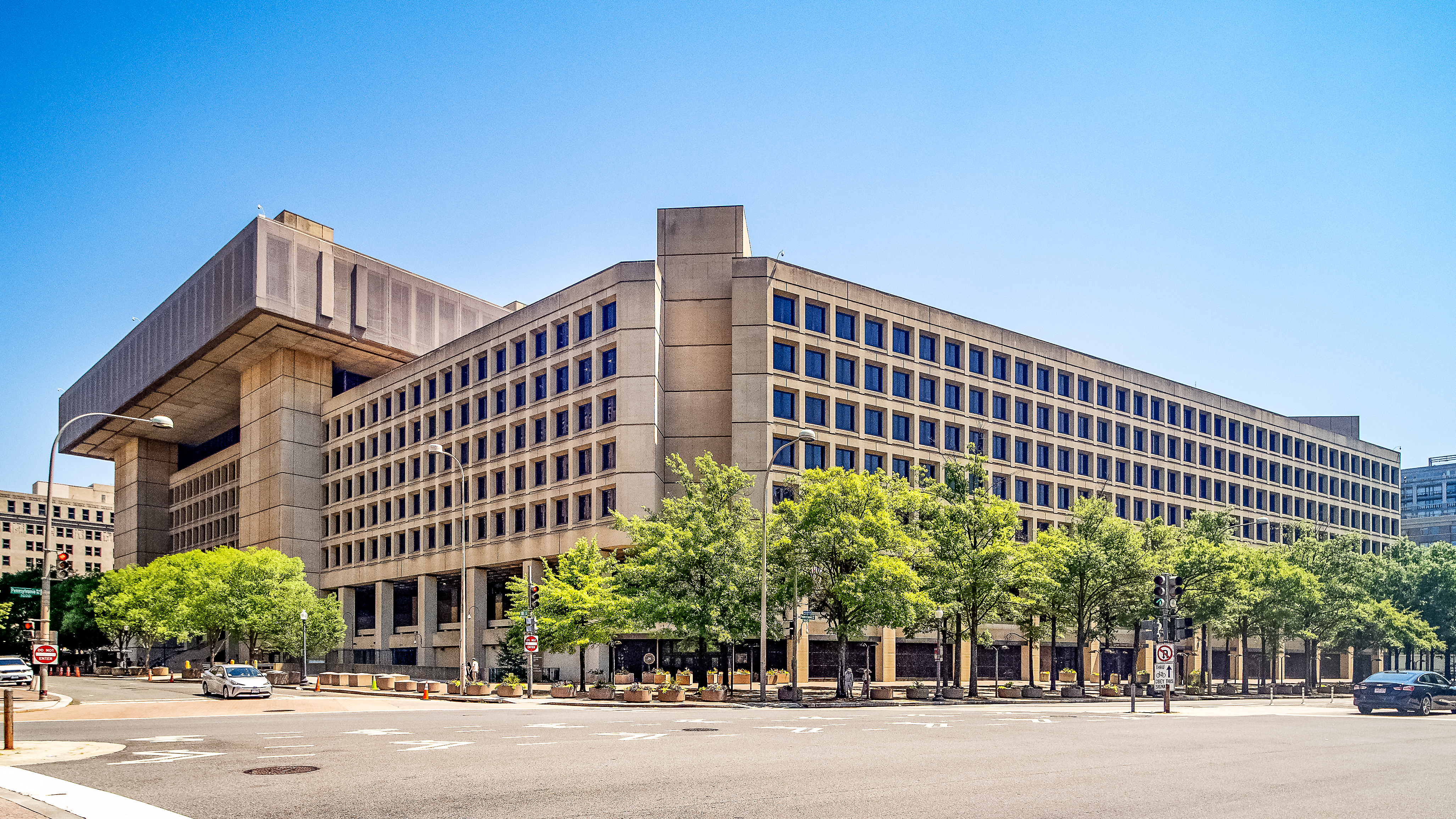 The FBI headquarters building in Washington, D.C. on an overcast day, with people walking past the entrance, photorealistic news photography