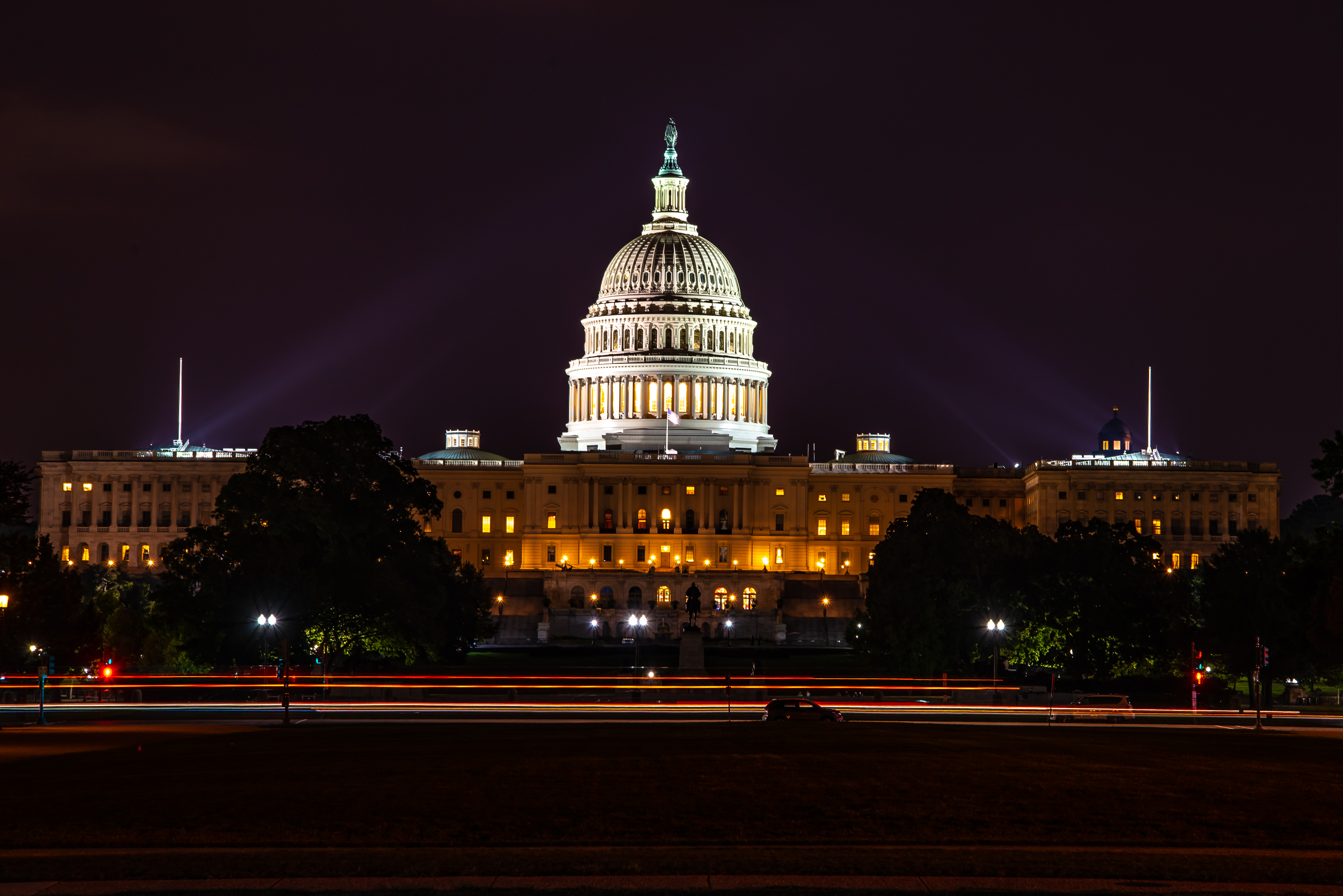 The United States Capitol building at dusk with lights on and a small number of pedestrians on the grounds, photographed from a distance, news photography style