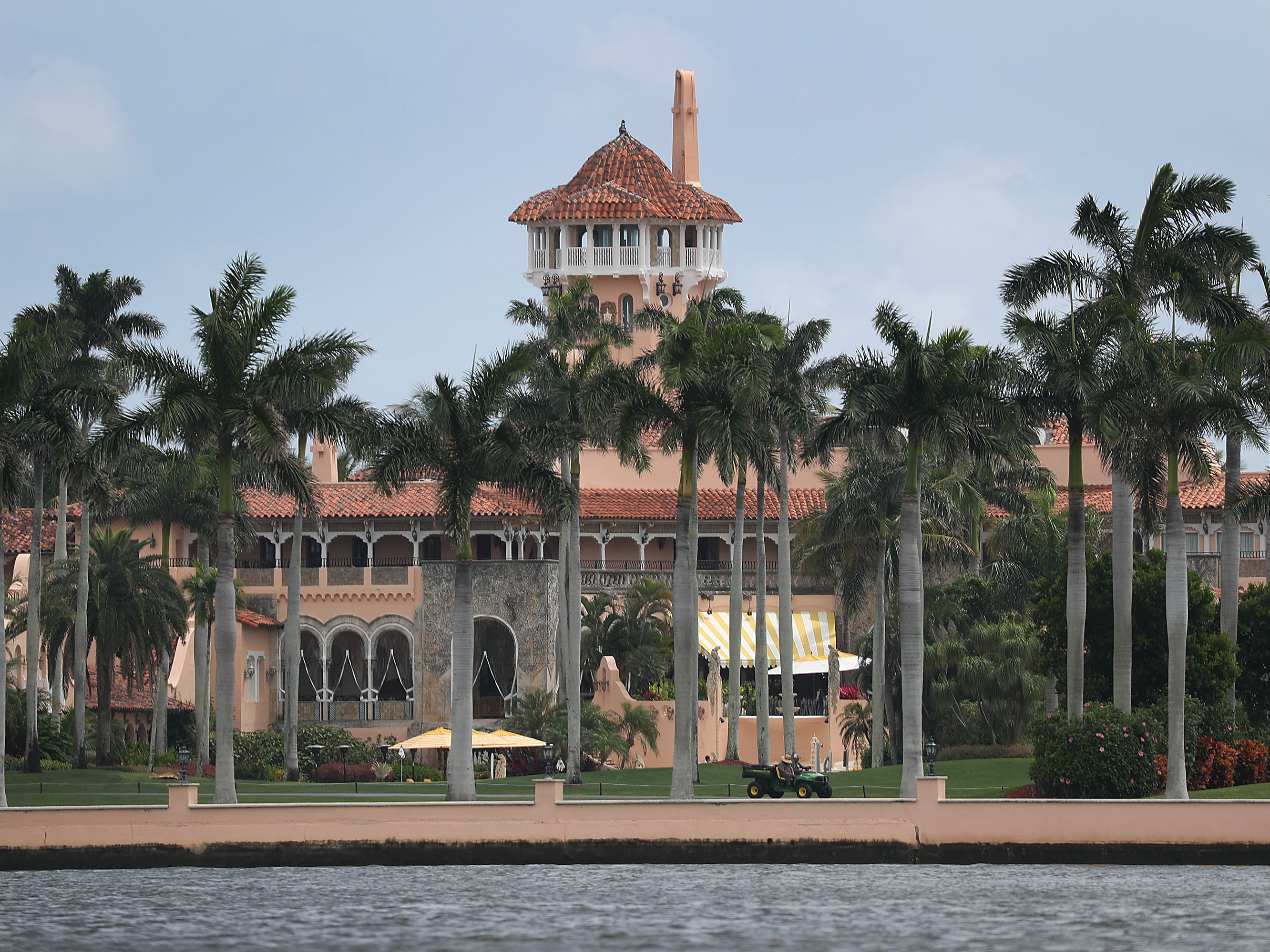 The exterior of Mar-a-Lago in Palm Beach, Florida, viewed from the street with palm trees and security fencing visible