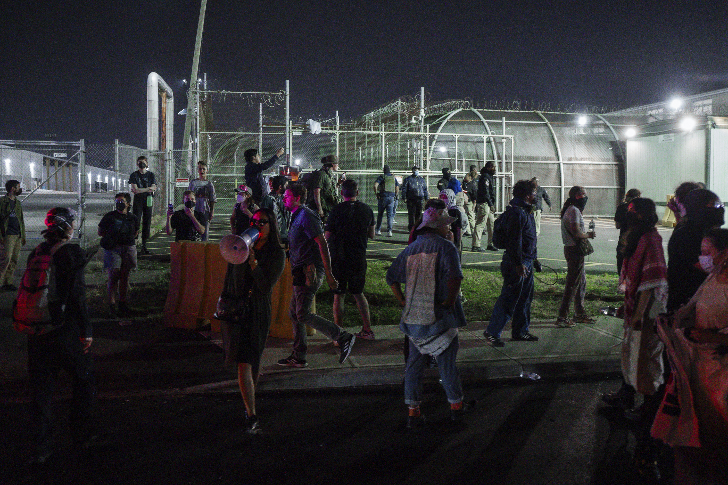 The exterior of an immigration detention facility at night in May 2026, with security lighting and fencing visible, news photography style