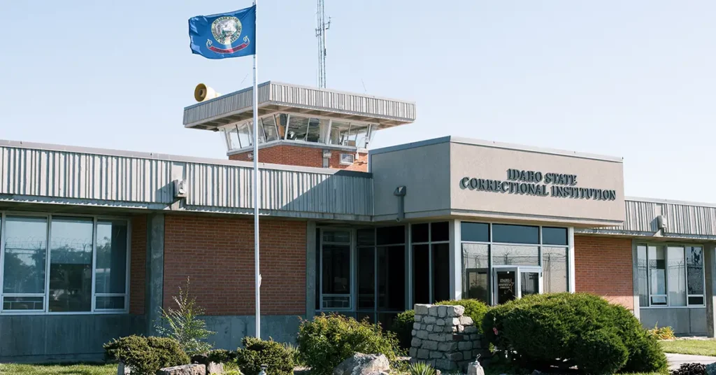 The exterior of the Idaho State Correctional Institution on a clear day, with a secure entrance and perimeter fencing visible, news photography style