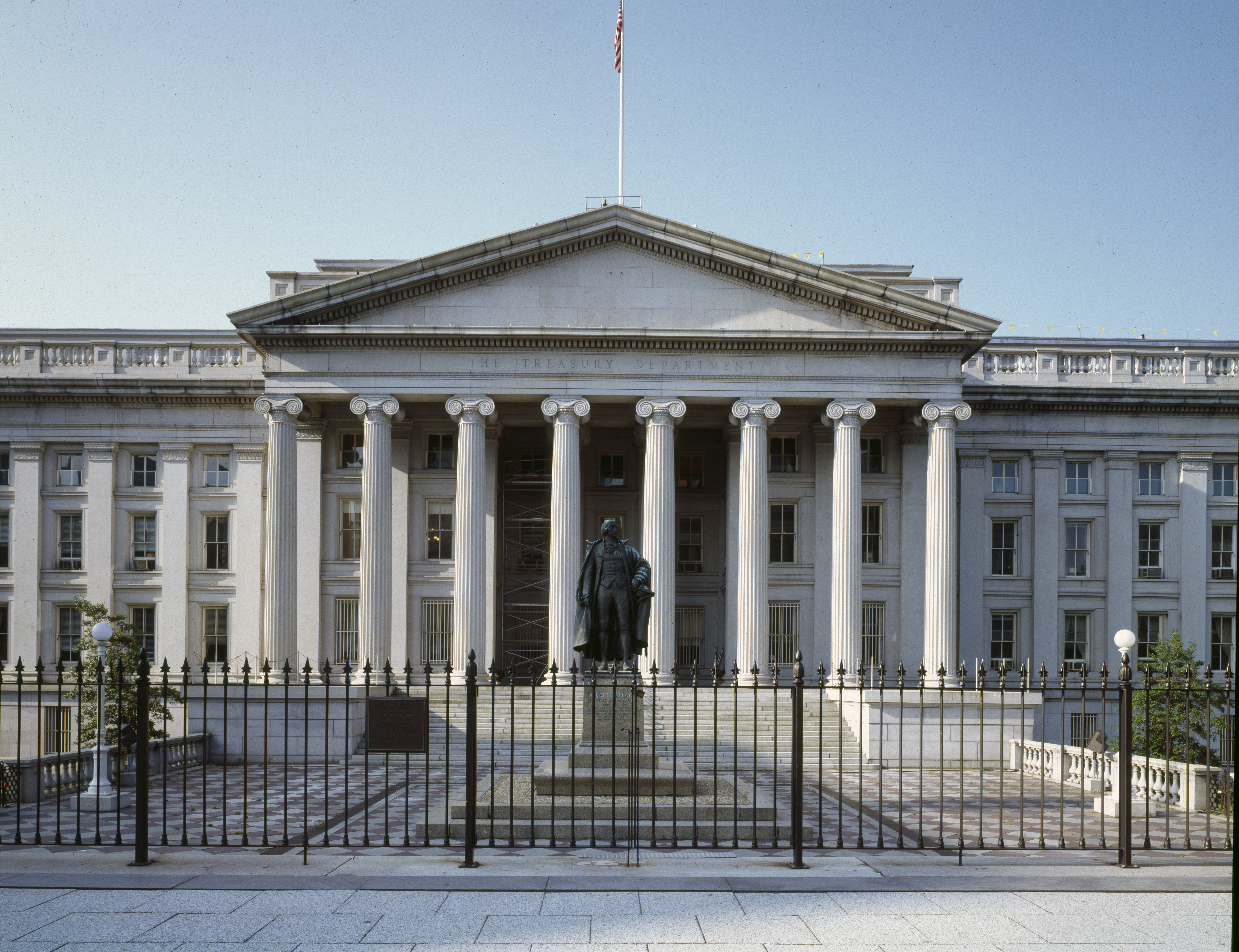 The exterior of the U.S. Treasury building in Washington, DC on a clear day, with columns and pedestrians in the foreground, news photography style