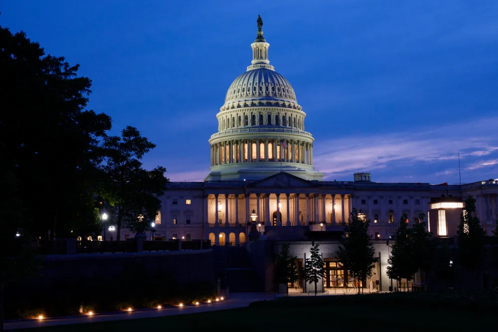 The exterior of the United States Capitol at dusk with warm lights glowing through windows and a few pedestrians on the grounds, news photography style