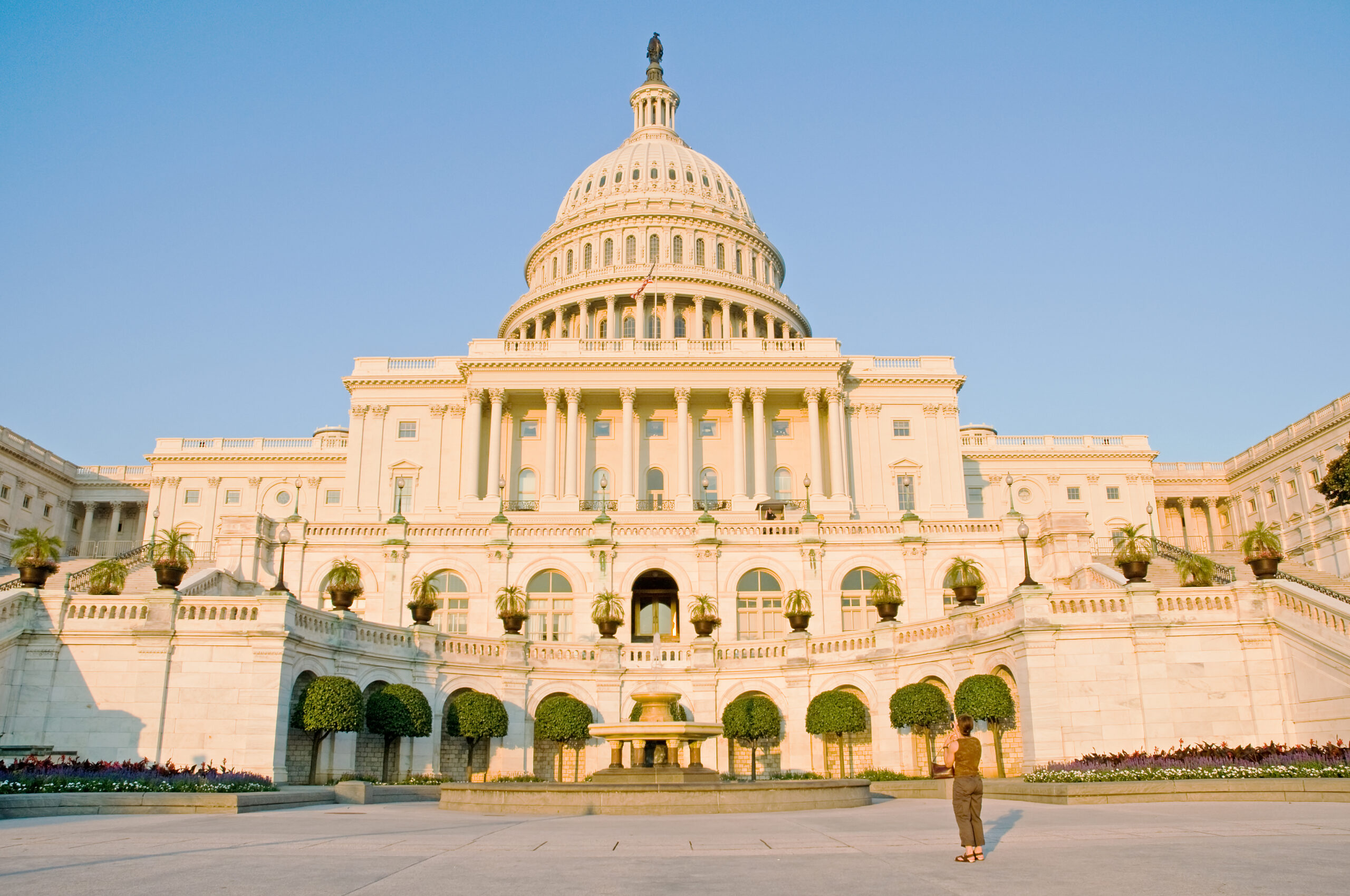 The exterior of the United States Capitol on the Senate side on a clear day, photographed from ground level with pedestrians in the distance, news photography style