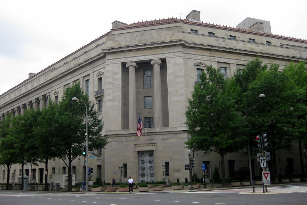 The exterior of the United States Department of Justice building in Washington, DC, photographed in daylight with people walking near the entrance, news photography style