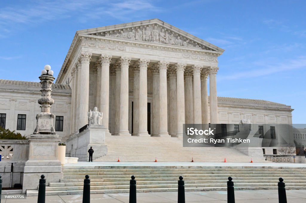 The front steps of the United States Supreme Court in Washington, DC on a clear day with a few people walking near the entrance, realistic news photography style