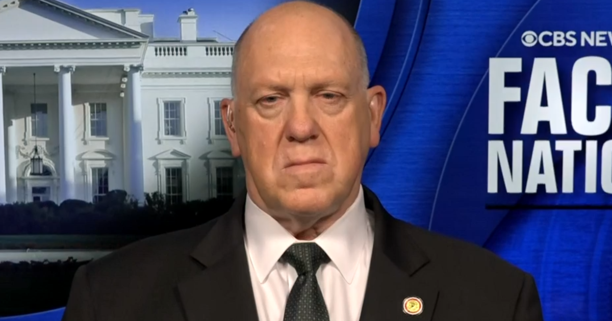 Tom Homan speaking during a televised studio interview, seated and facing a news host, with neutral background lighting, news photography style