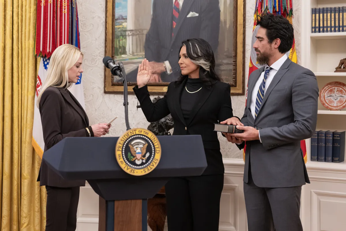 Tulsi Gabbard shaking hands with President Donald Trump in the Oval Office after a swearing-in moment, with flags and the Resolute Desk visible, news photography style