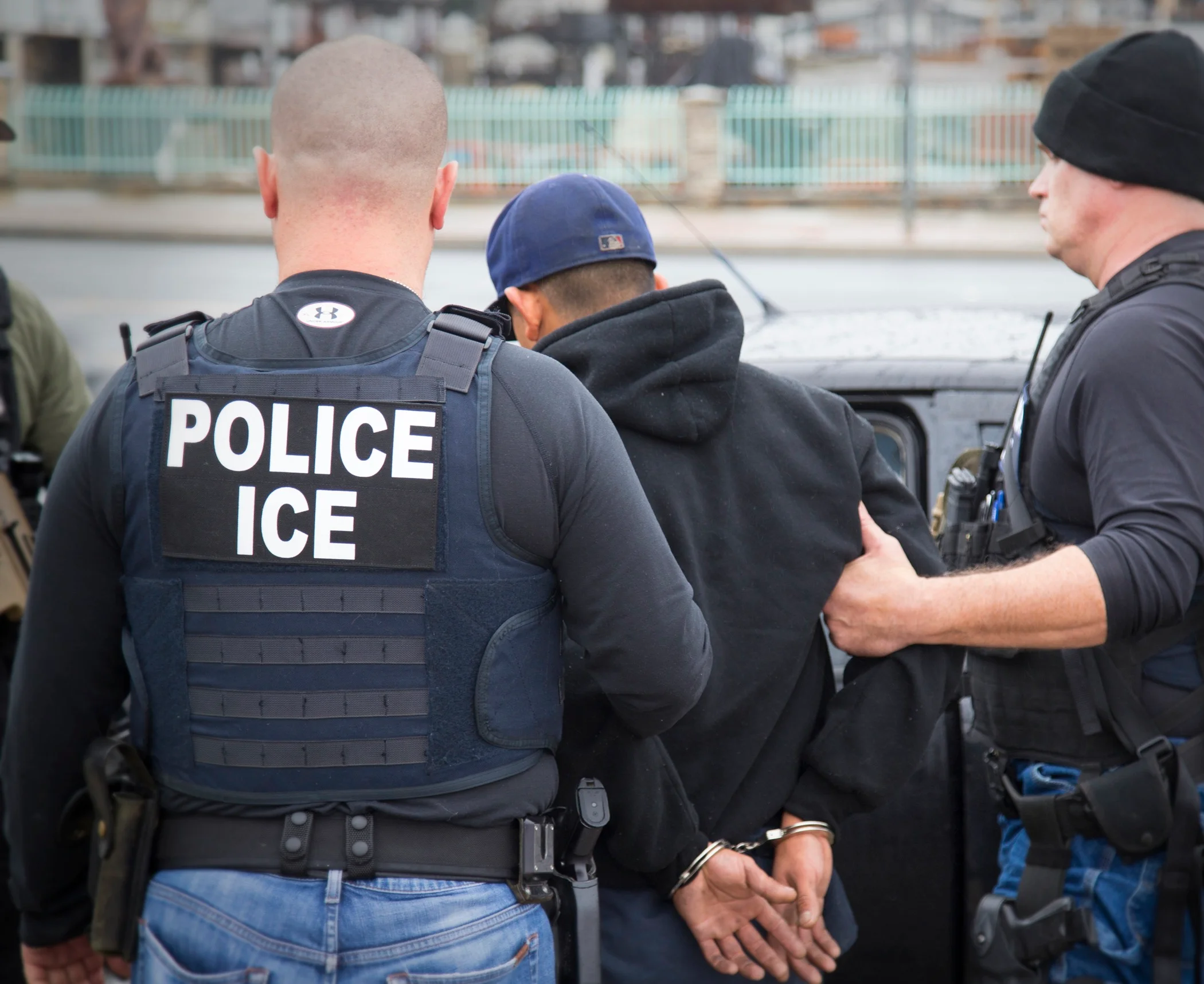 Two ICE agents in tactical vests walking with a detained individual in an urban setting, photographed from a distance in a candid news style