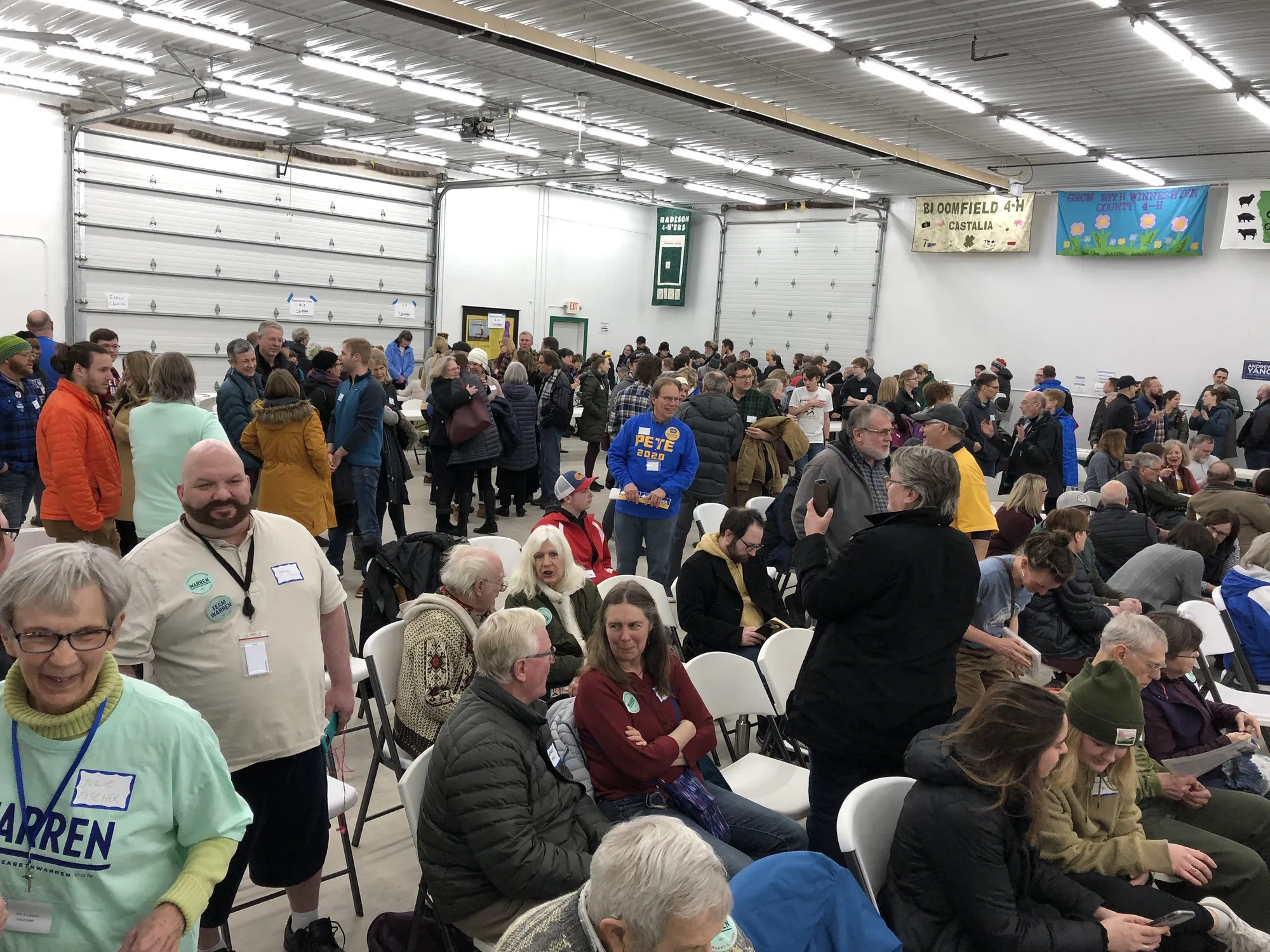 Voters gathered inside a school gym during a caucus precinct meeting, standing in clusters to show support for different candidates, documentary news photo style