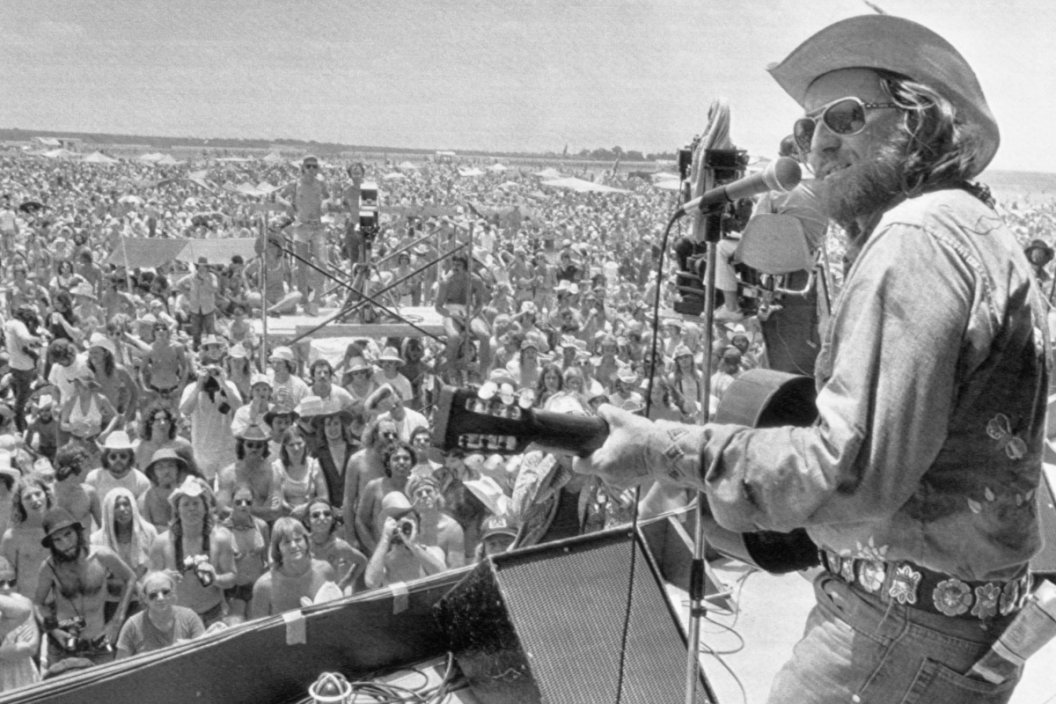 Willie Nelson performing onstage at his Fourth of July Picnic in Austin, with concert lighting and instruments visible